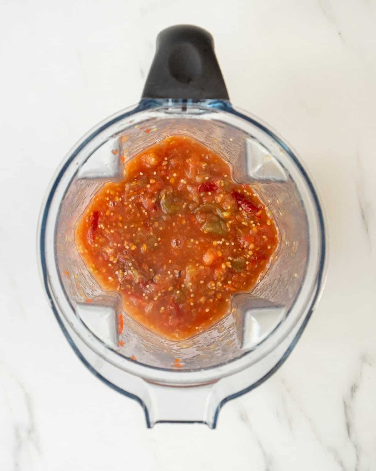 A blender filled with blended dry chilies, tomatillos, tomatoes, and garlic cloves on a white countertop. 