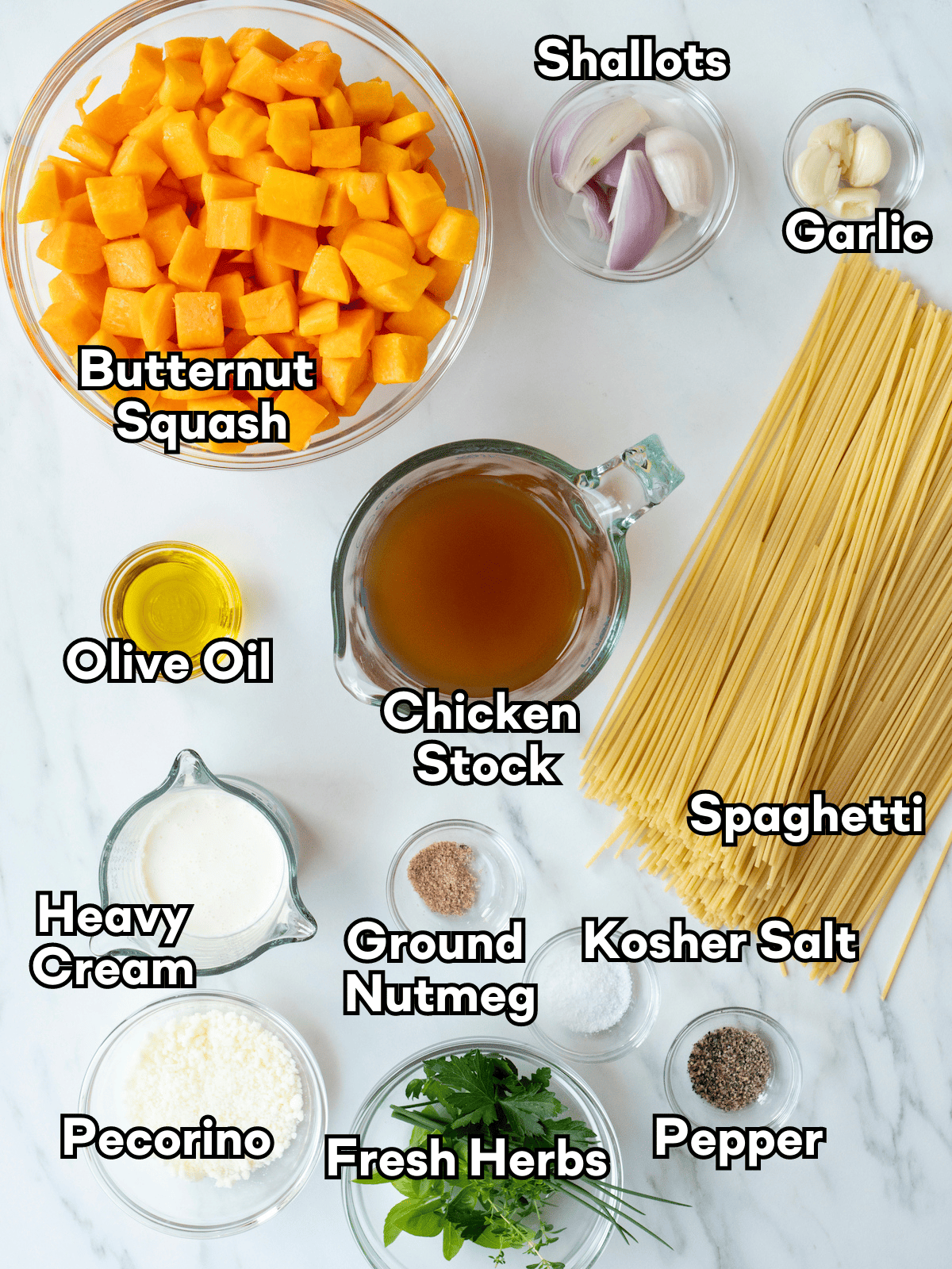 An overhead shot of all the ingredients needed to make this recipe.  Ingredients consist of butternut squash, shallots, garlic, olive oil, chicken stock, spaghetti, heavy cream, ground nutmeg, kosher salt, pecorino, fresh herbs, and pepper.  