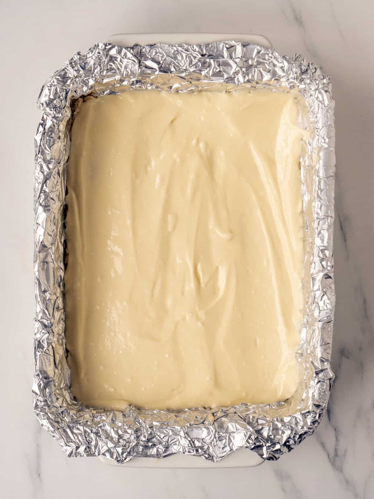 A 9x13 baking pan lined with tin foil with cookie dough pressed into the bottom of the pan that has a layer of the cheesecake mixture evenly spread on top of it.