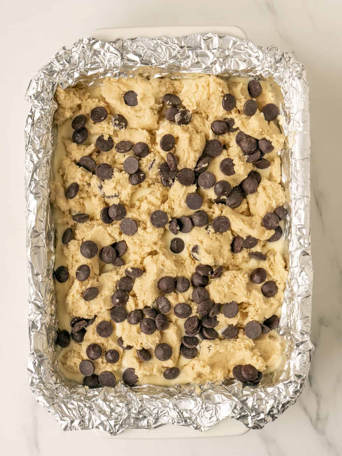 A 9x13 baking pan lined with tin foil with crumbled cookie dough sprinkled all over the cookie dough and cream cheese mixture layers below, and a final layer of chocolate chips sprinkled on top.