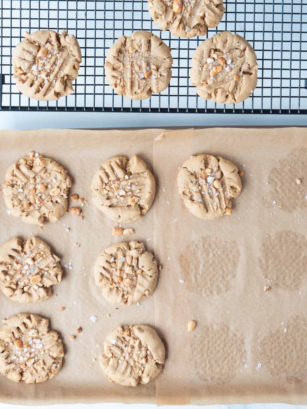 A wire rack along with a parchment-lined baking sheet, both with peanut butter cookies and a few crumbs and peanuts on the parchment.