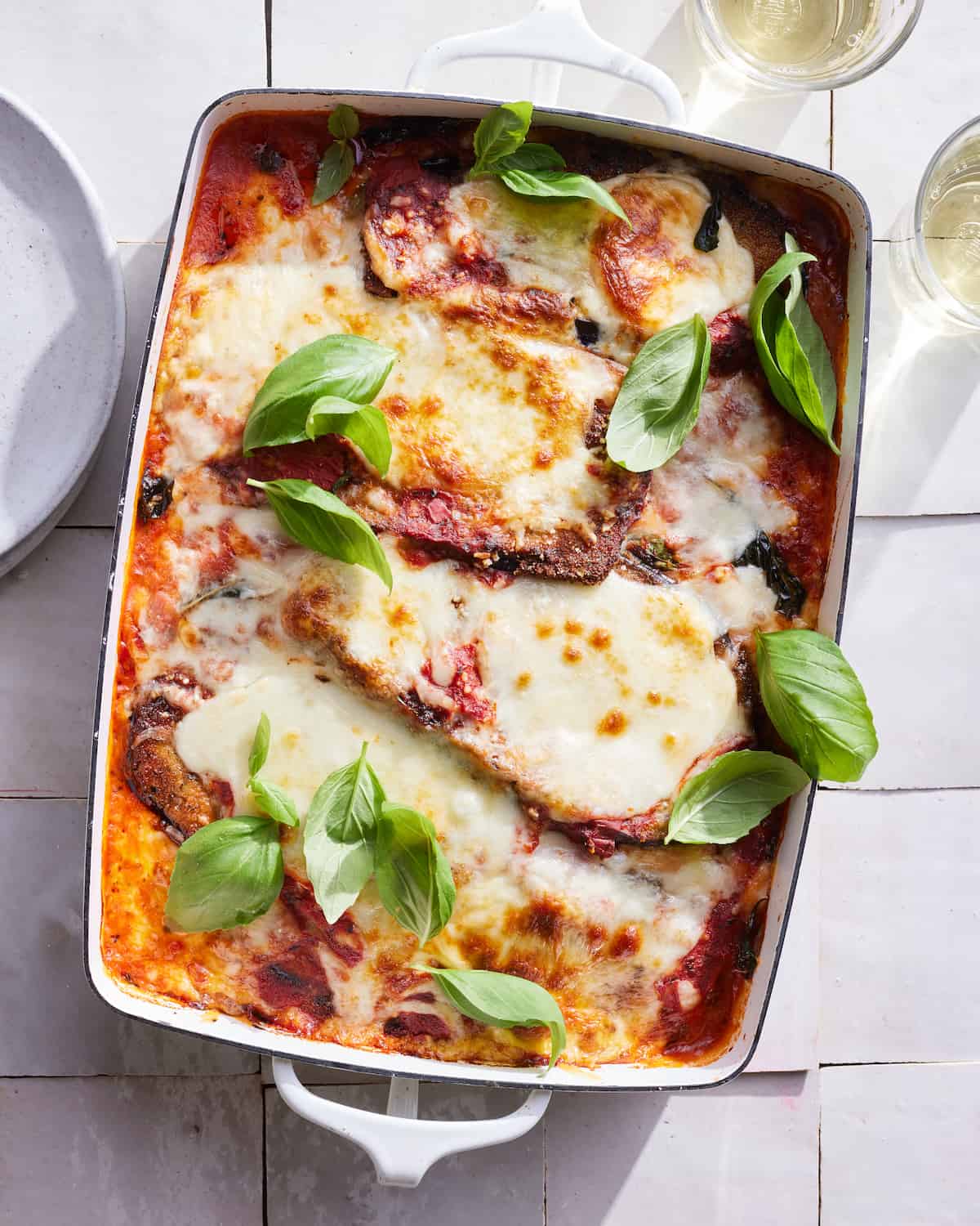 Prepared eggplant parmesan in ceramic baking dish sitting on a countertop surrounded by a ceramic plate and two drinking glasses.