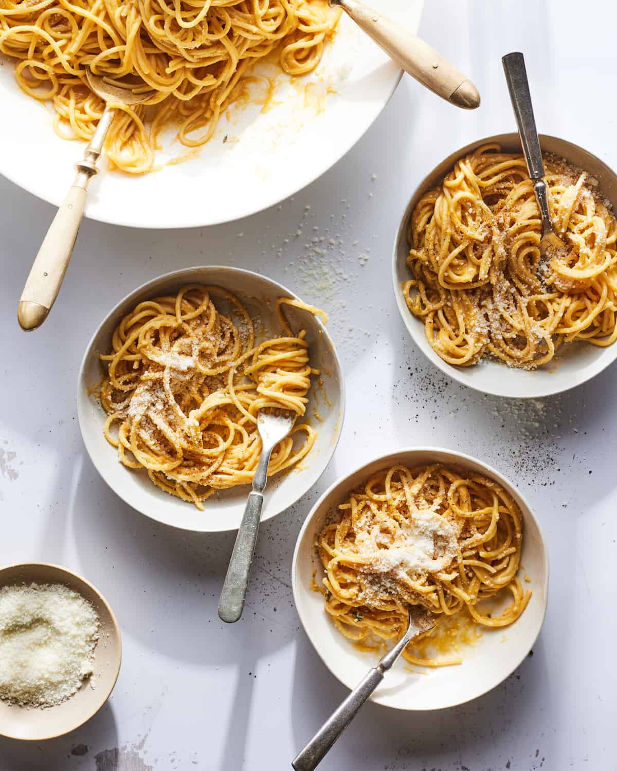 Three individual bowls of butternut squash pasta next to a large serving bowl of pasta and a small bowl of parmesan cheese.  