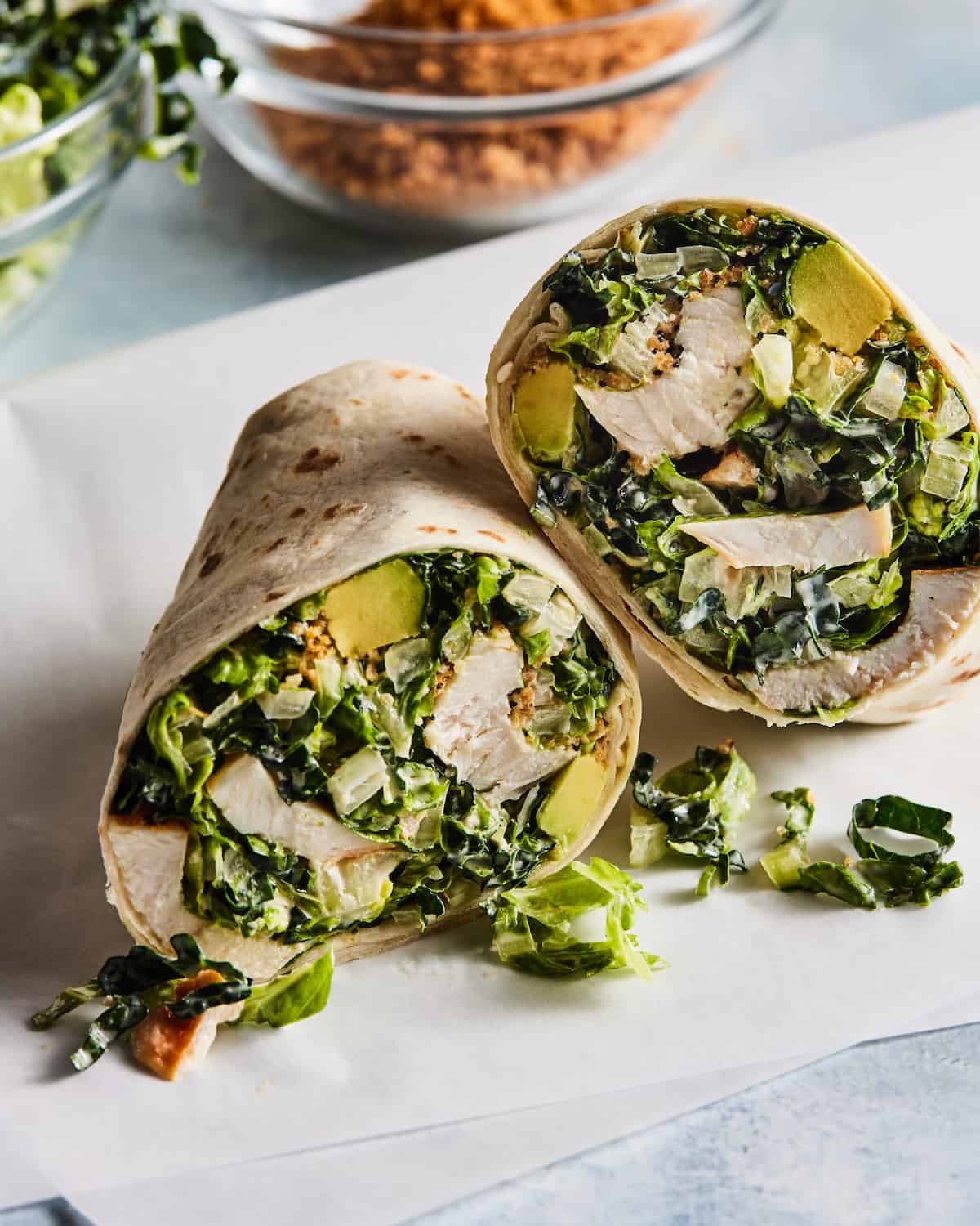 A Chicken Caesar Wrap split in half sitting on some parchment paper with a bowl of breadcrumbs and kale in the background.  