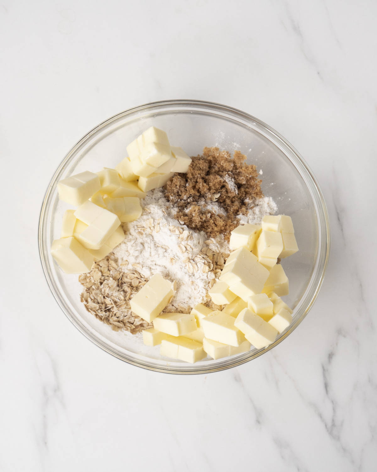 Clear mixing bowl with oats, flour, dark brown sugar, and cubed butter on a white countertop.