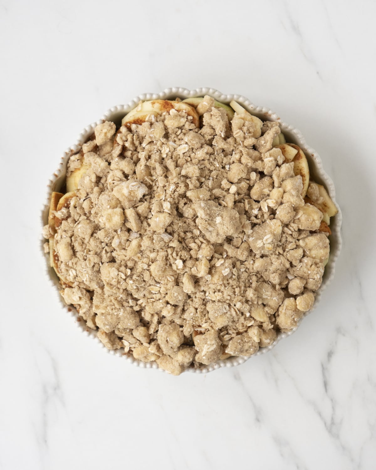 Ceramic baking dish of sliced apples and cinnamon topped with streusel topping mixture on a white countertop.