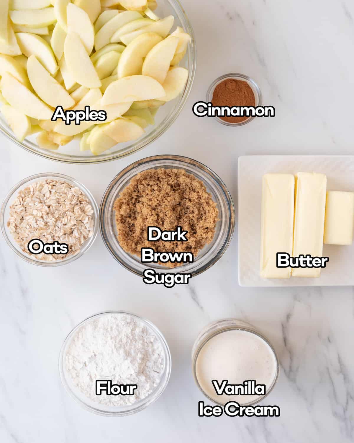 Ingredients in individual clear bowls such as sliced apples, cinnamon, oats, dark brown sugar, butter, flour, and vanilla ice cream on a white countertop.