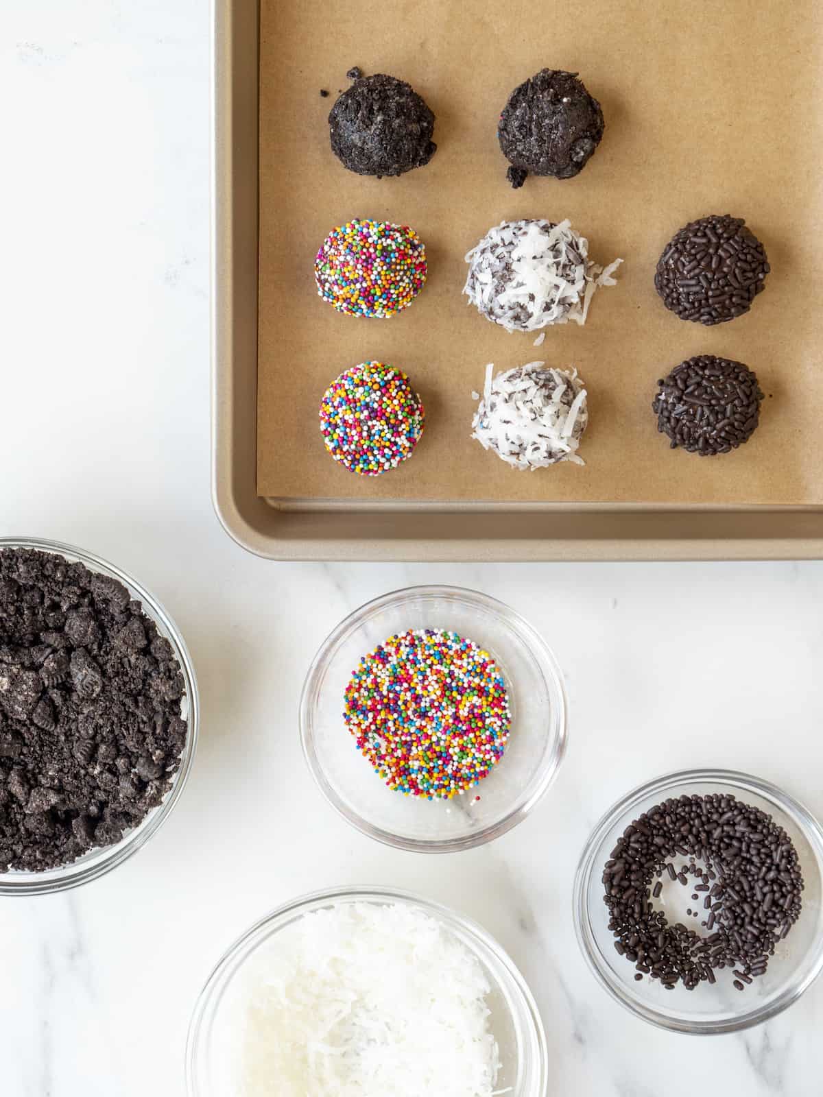 4 small bowls with toppings consisting oreos, rainbow sprinkles, chocolate sprinkles and shredded coconut and a parchment lined baking sheet of brigadeiros coated, each in one of these.