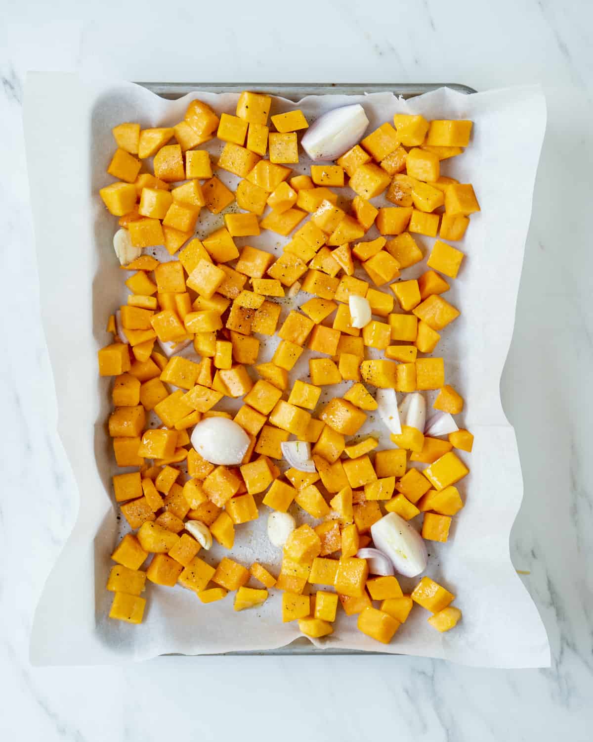 A sheet pan lined with parchment paper of butternut squash, shallots, garlic, and drizzled olive oil, seasoned with salt and pepper.