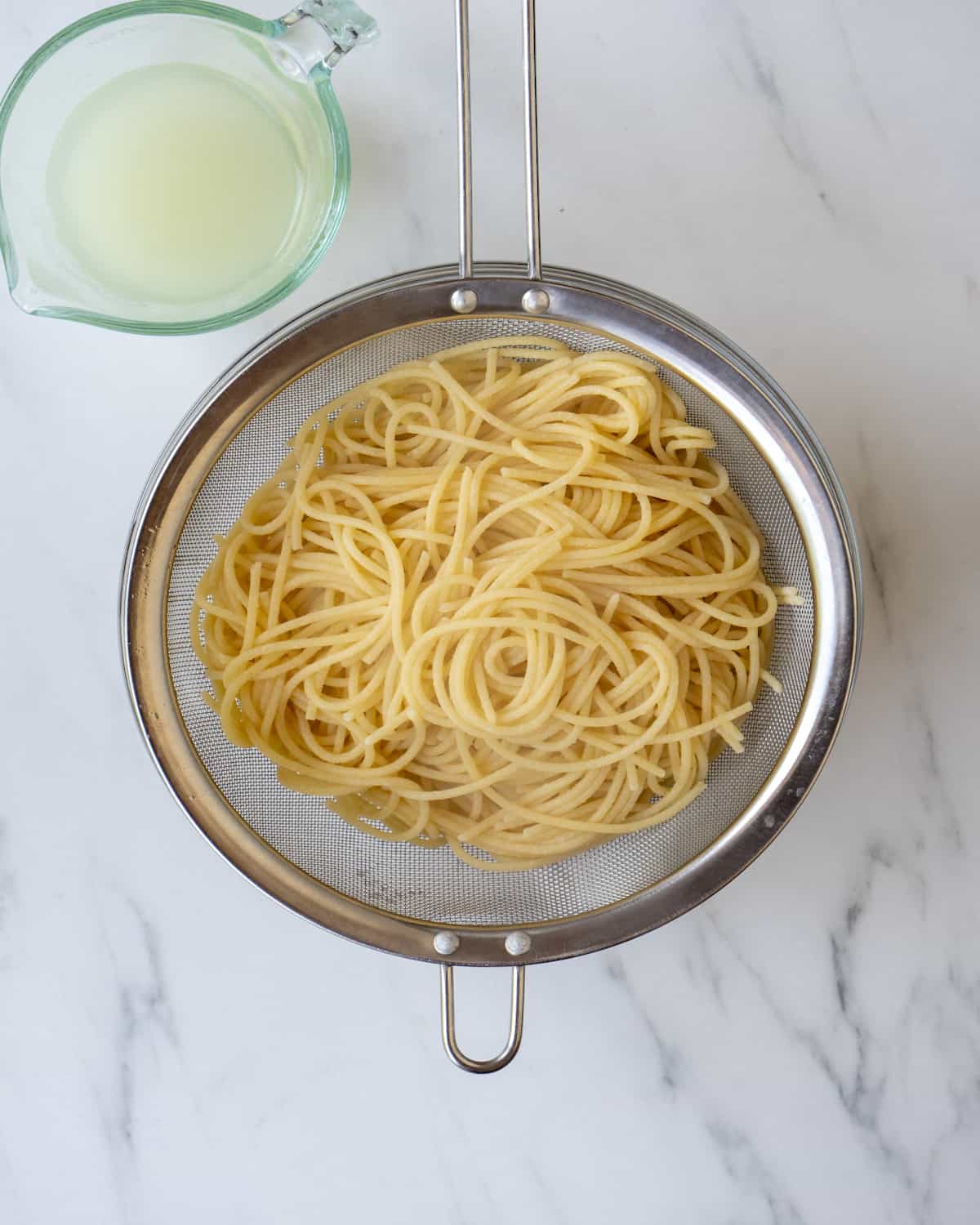 A metal strainer with cooked spaghetti noodles and a measuring cup of reserved pasta water.