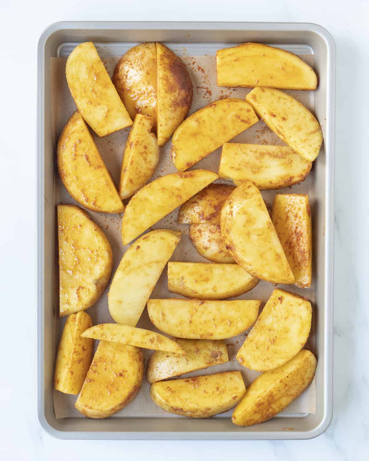 Potato wedges on a metal sheet pan lined with parchment paper on a white countertop.