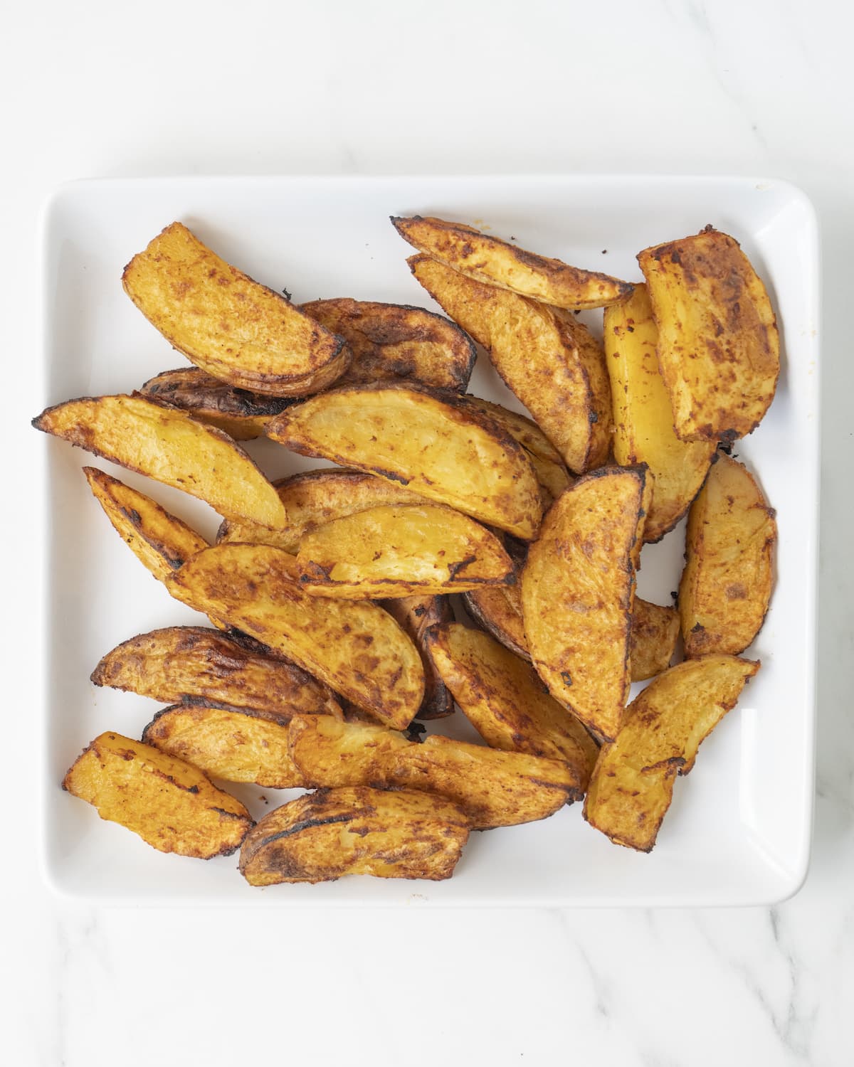 Cooked potato wedges on a white oven-safe ceramic plate on a white countertop.