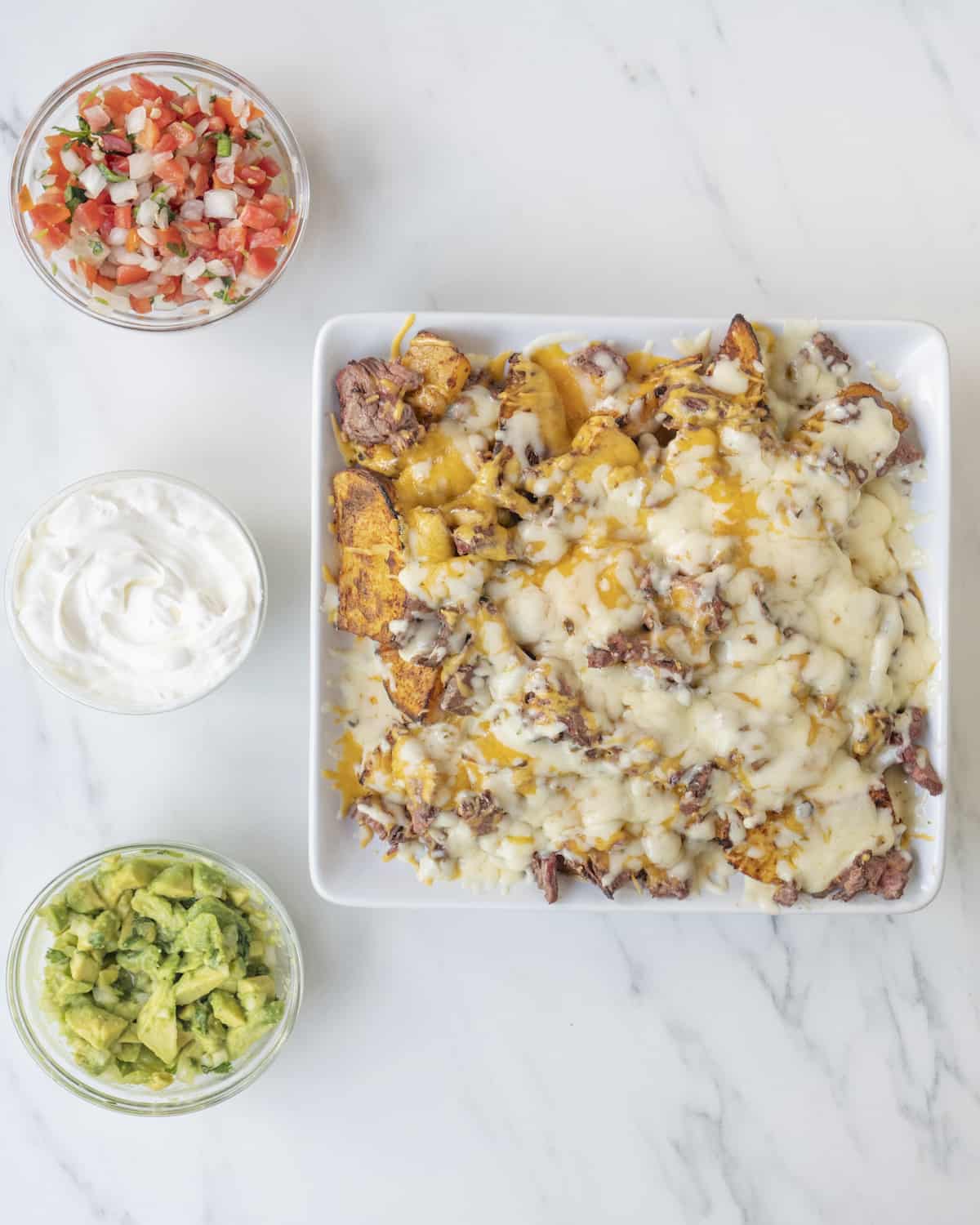 Assembled carne asada nacho fries on a white dish with three clear bowls of topping ingredients such as pico de gallo, sour cream, and guacamole on a white countertop.