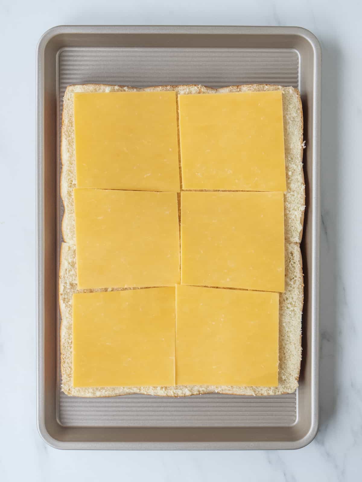 A baking sheet with a 3x4 grid of bottom half of slider buns topped with slices of cheddar cheese.