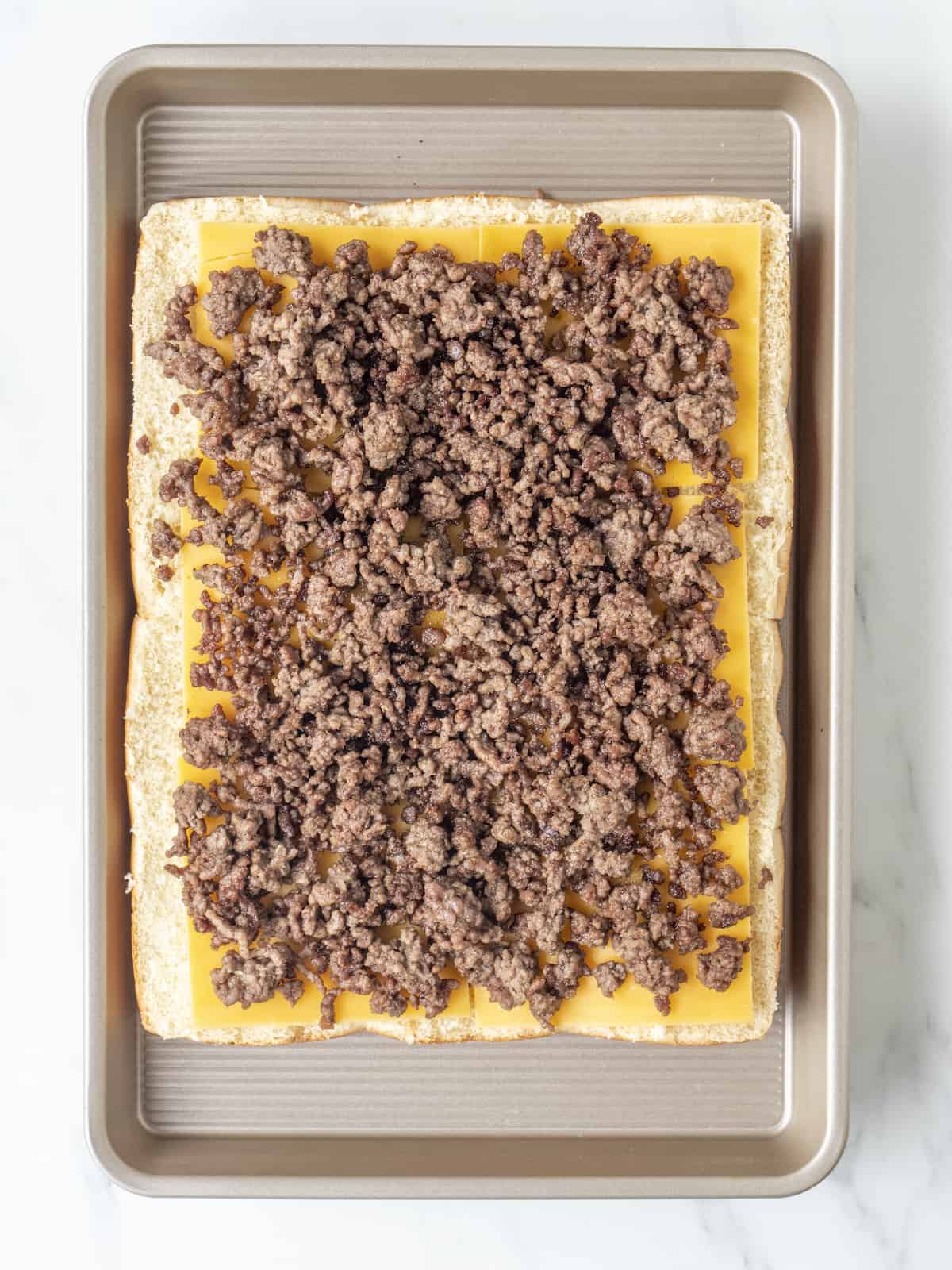 A baking sheet with a 3x4 grid of bottom half of slider buns topped with slices of cheddar cheese and ground beef.