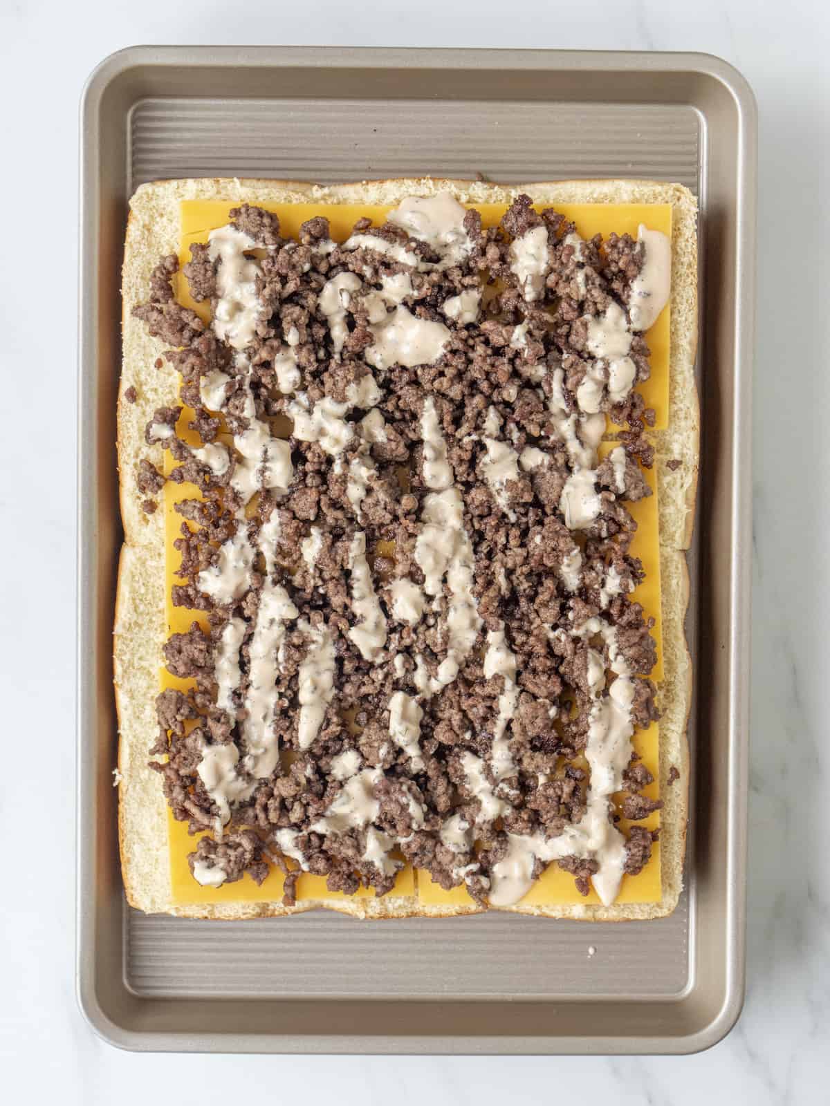 A baking sheet with a 3x4 grid of bottom half of slider buns topped with slices of cheddar cheese, ground beef and sauce.