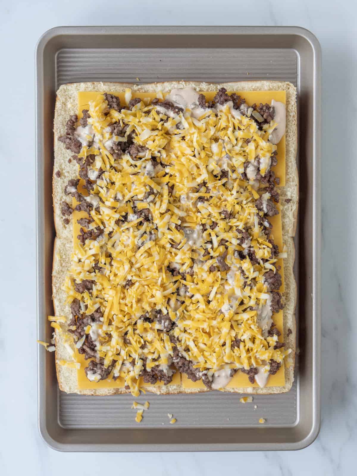 A baking sheet with a 3x4 grid of bottom half of slider buns topped with slices of cheddar cheese, ground beef, sauce and shredded cheese.