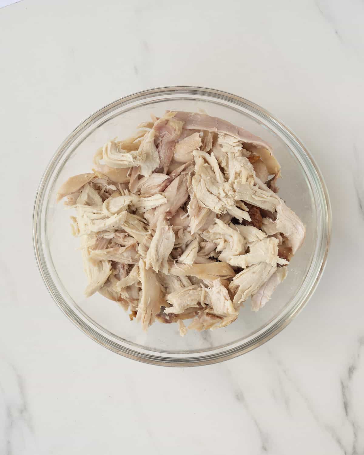 Shredded rotisserie chicken in a clear bowl on top of a white countertop.