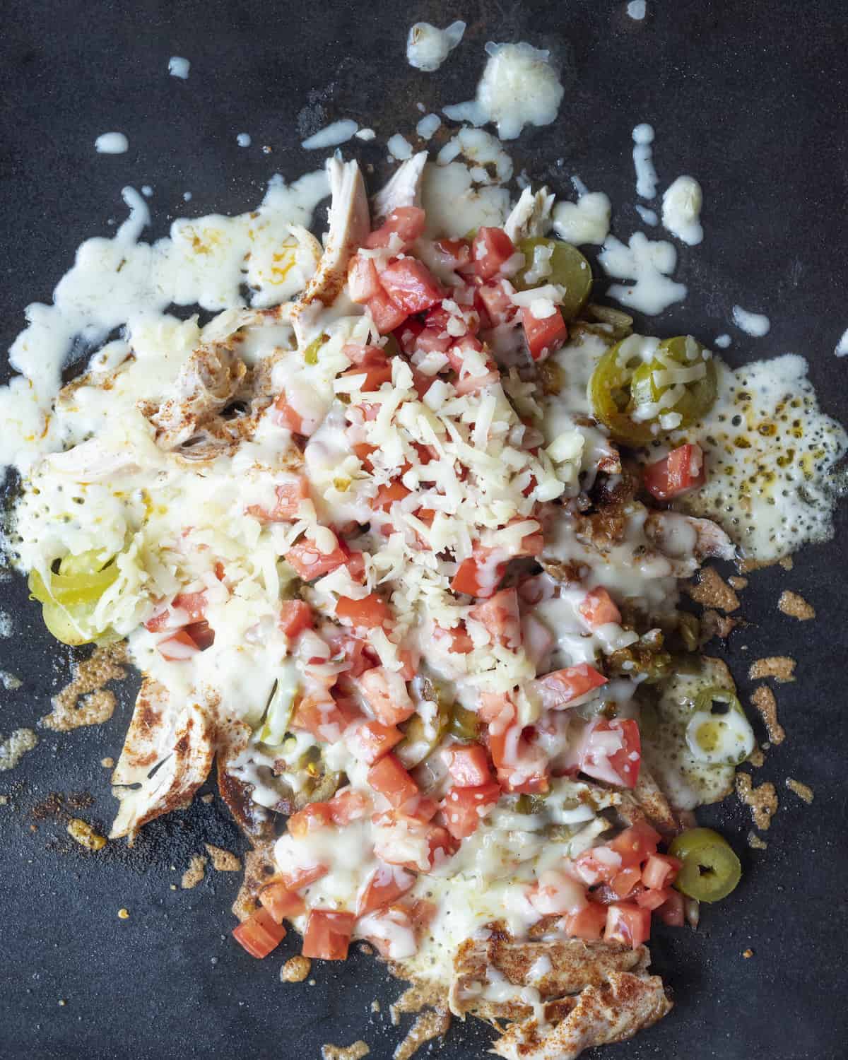 Shredded rotisserie chicken on a griddle mixed with chopped green chiles and pickled jalapeños and taco seasoning topped with shredded cheese and diced tomatoes.