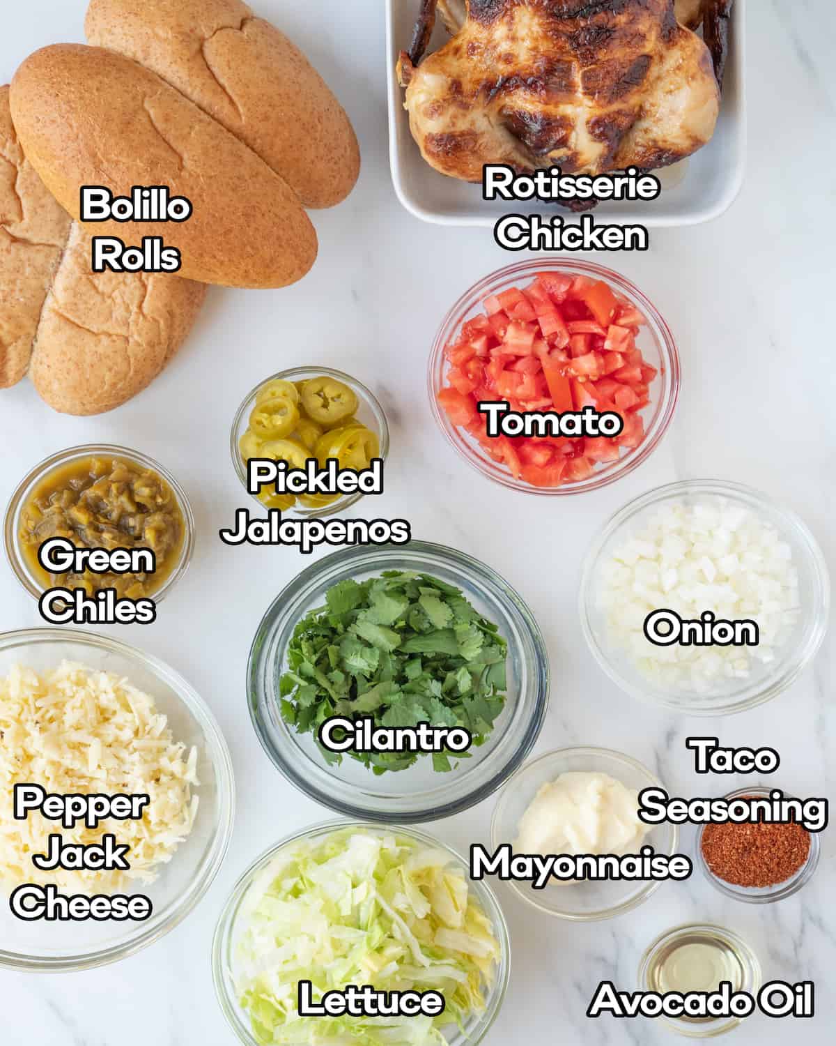 Every ingredient you need to make this recipe such as bolillo rolls, rotisserie chicken, green chiles, pickled jalapeños, tomatoes, onion, cilantro leaves, pepper jack cheese, mayonnaise, taco seasoning, lettuce, and avocado oil in individual bowls on a white countertop. 