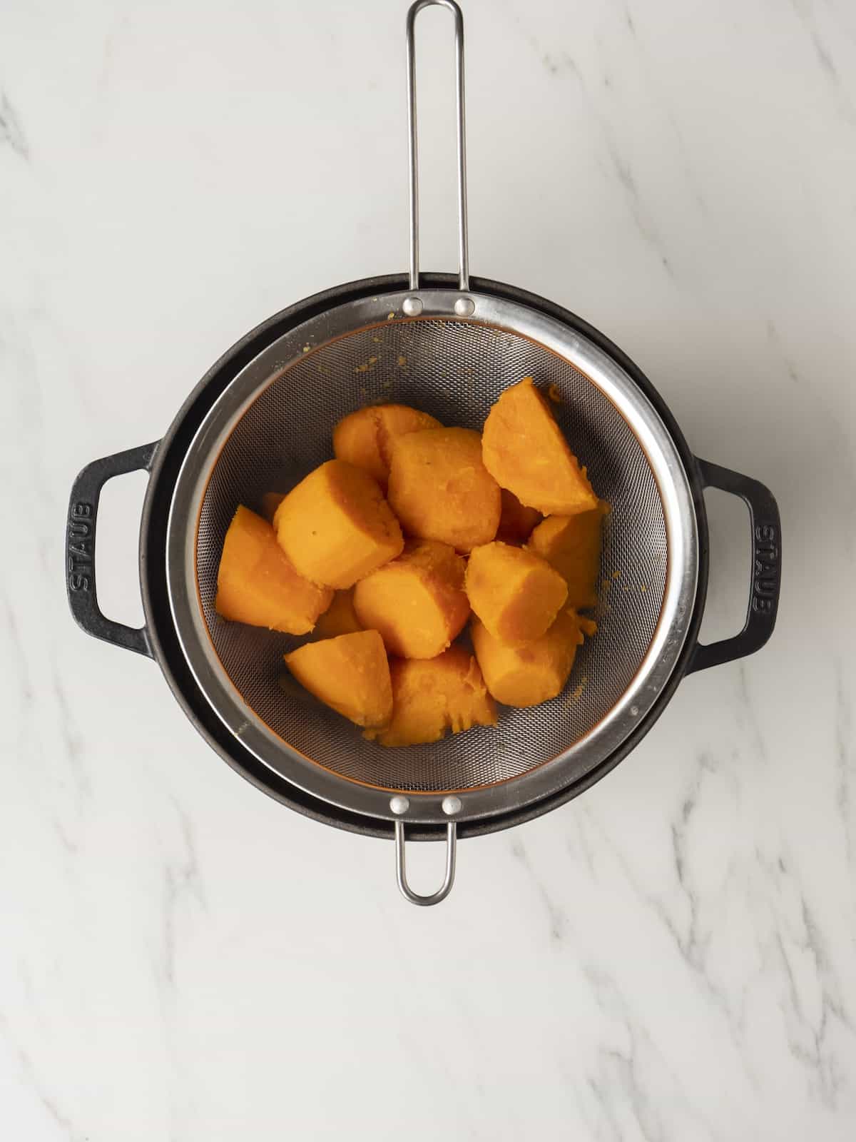 A strainer with boiled chunks of sweet potatoes, in a pot.