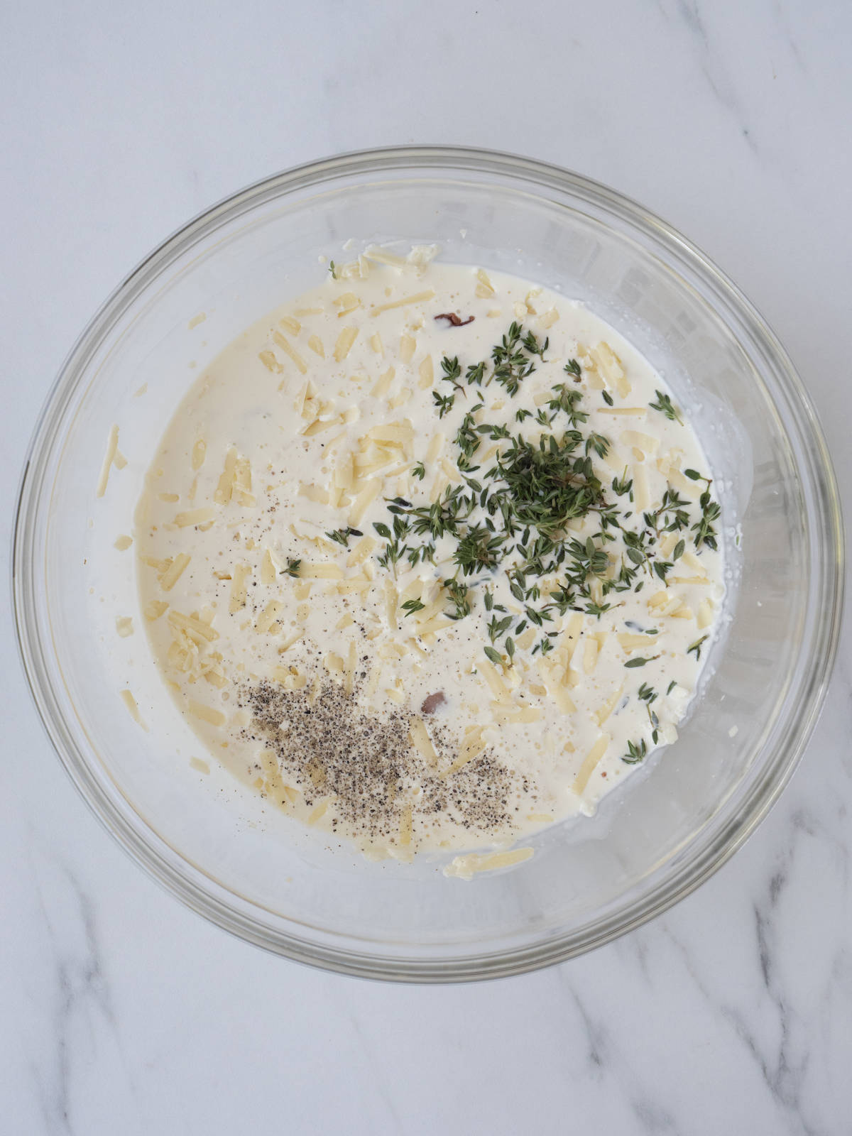 A glass mixing bowl with cheese, pancetta, thyme, cream and garlic, seasoned with salt and pepper.