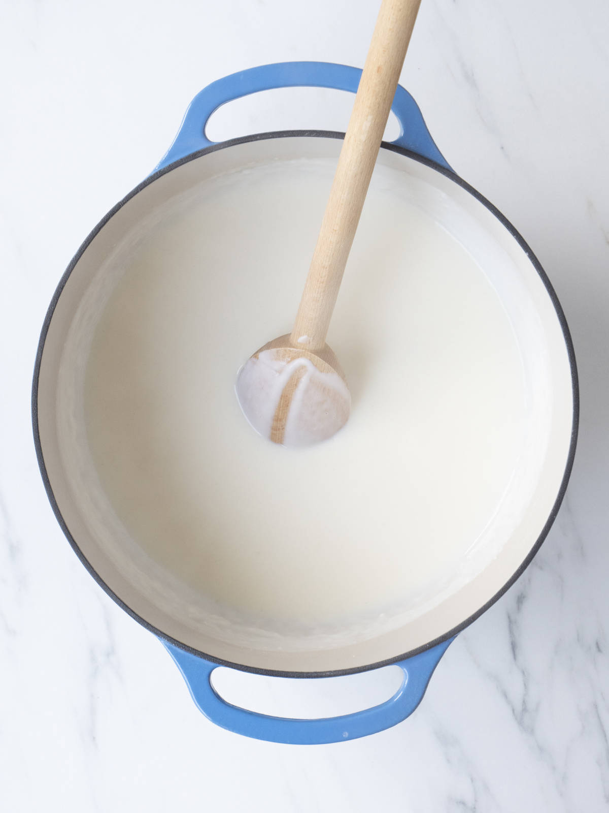 A blue dutch oven with a white sauce, comprising roux and milk with a spoon to mix.