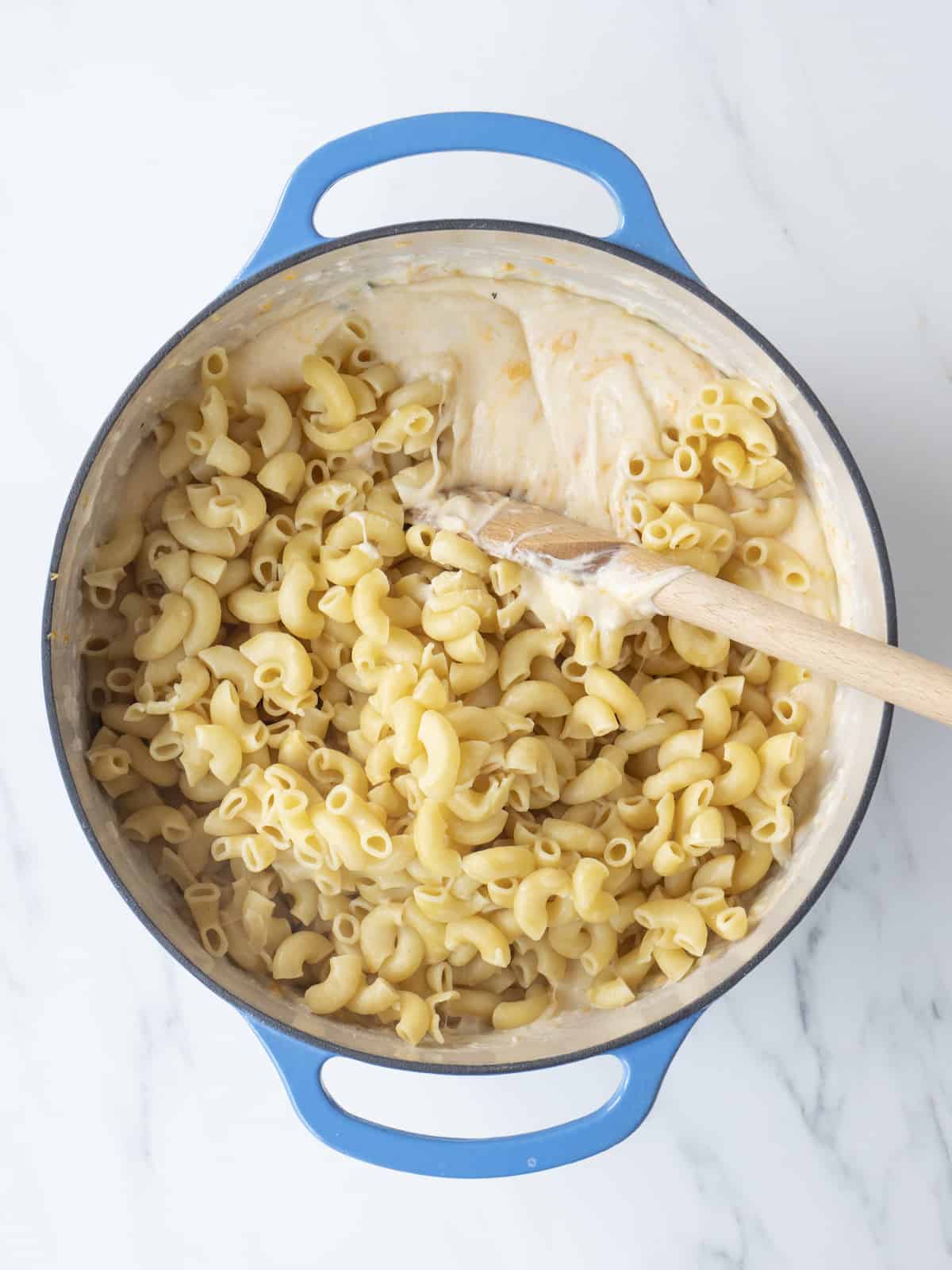 A blue dutch oven with macaroni being stirred into cheese sauce.