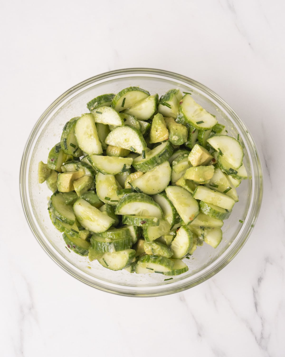 Diced cucumber and diced avocado combined with oil and lemon and seasoning mixture in a clear white bowl on top of a white countertop.