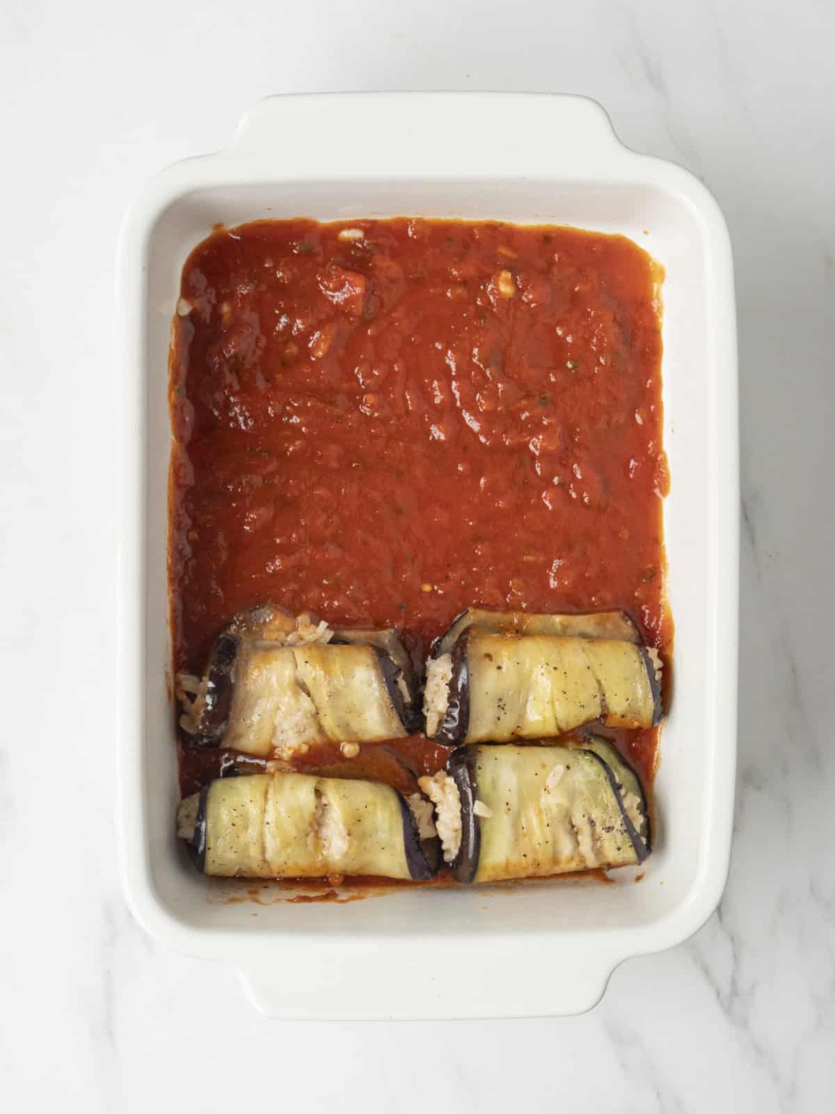 A rectangular white baking dish with marinara sauce in the base with 4 eggplant slices rolled up stuffed with parmesan risotto.