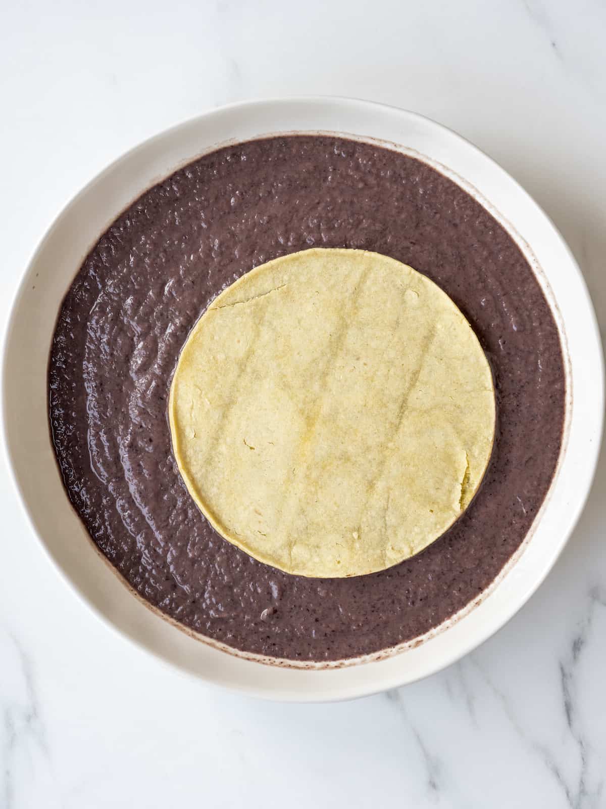 A corn tortilla placed on top of a white bowl with black bean puree.