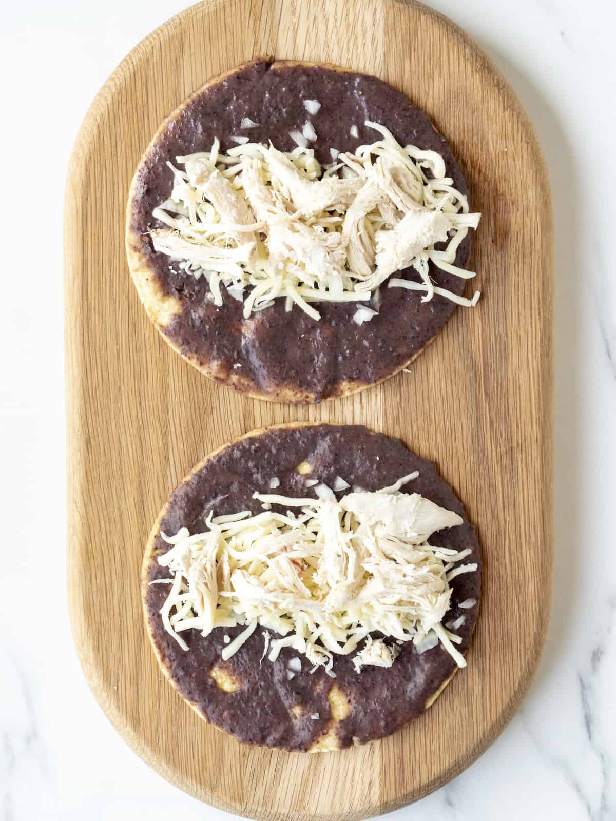 A wooden board with two corn tortillas with black bean puree, shredded chicken and shredded cheese.