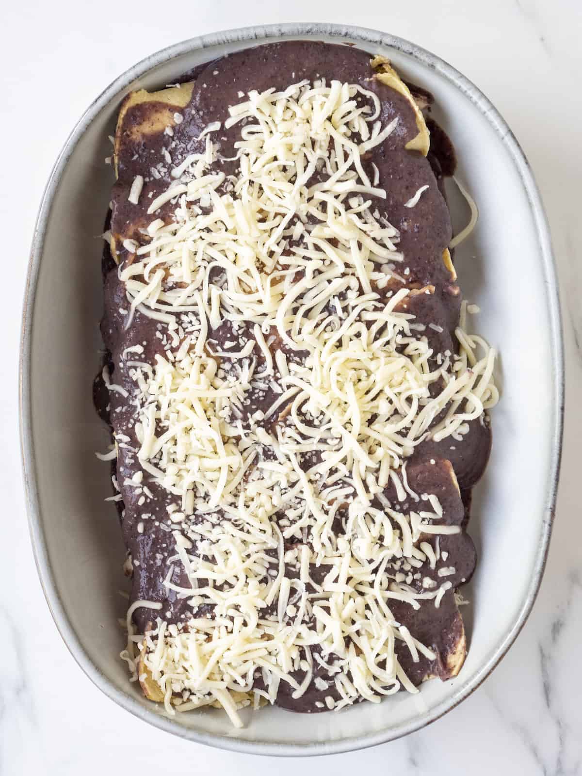 A grey shallow baking dish with rolled up tortillas stuffed with black bean puree, chicken and cheese, layered with more black bean puree and shredded cheese.