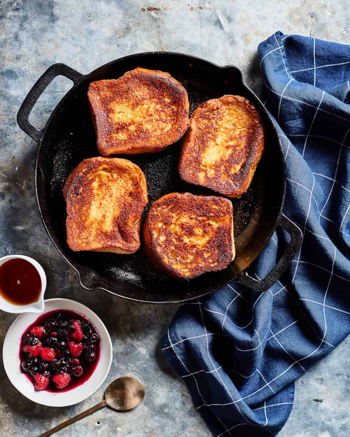 Caramelized French Toast in a skillet placed on a blue kitchen towel with a side serving of berry compote and maple syrup.