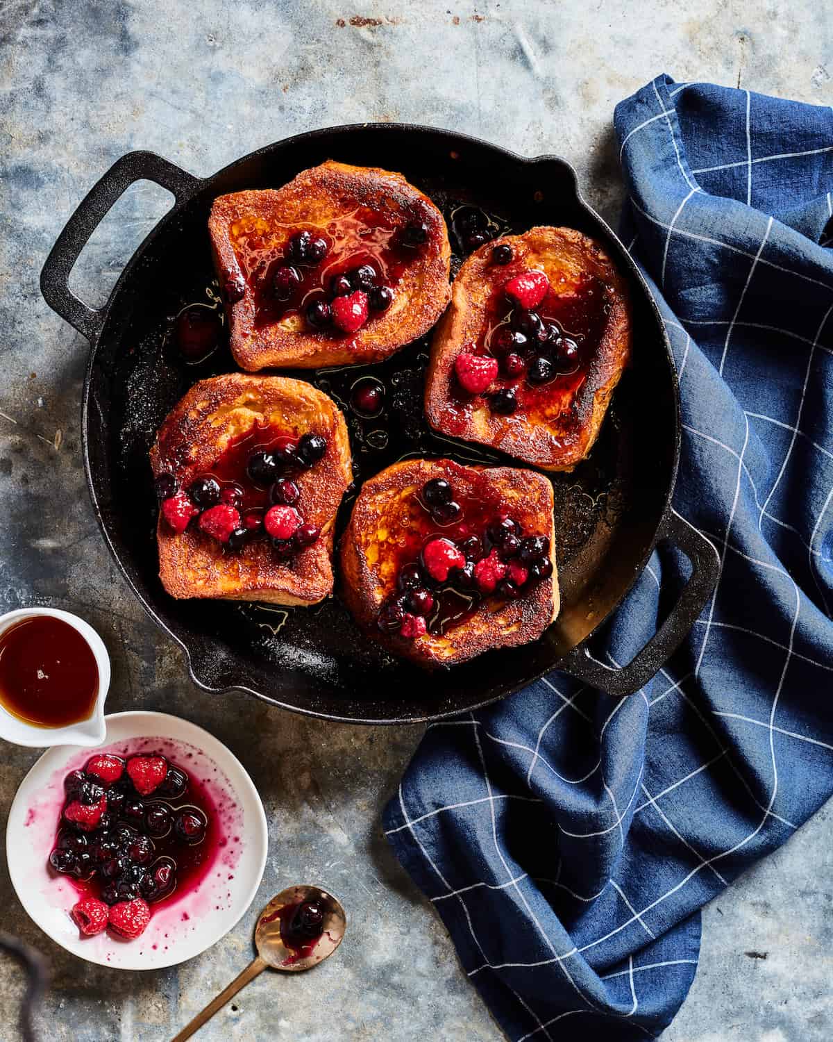 Caramelized French Toast with Berry Compote in a skillet placed on a blue kitchen towel with a side serving of berry compote and maple syrup.