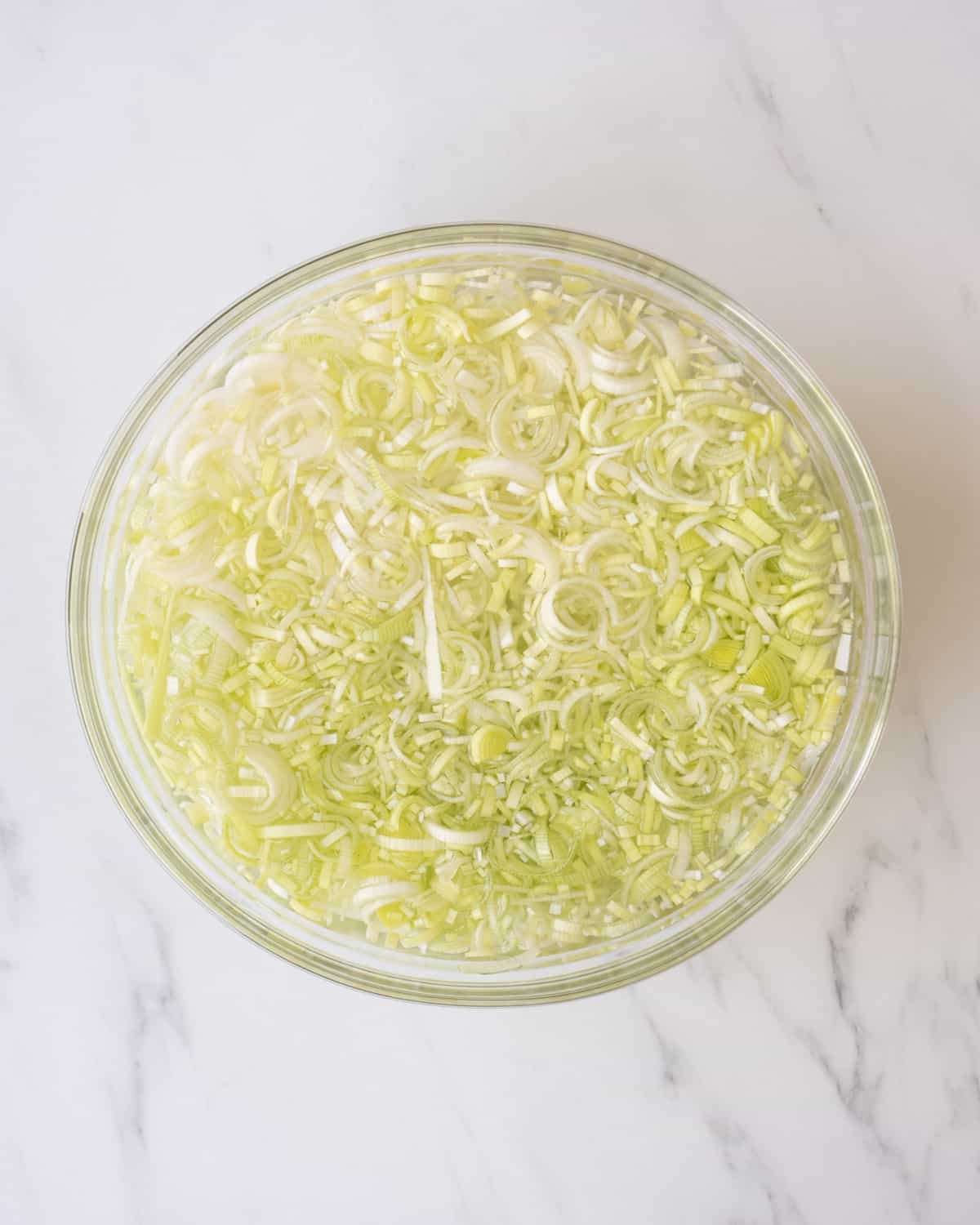 A clear bowl of chopped leeks in cold water on a white countertop.