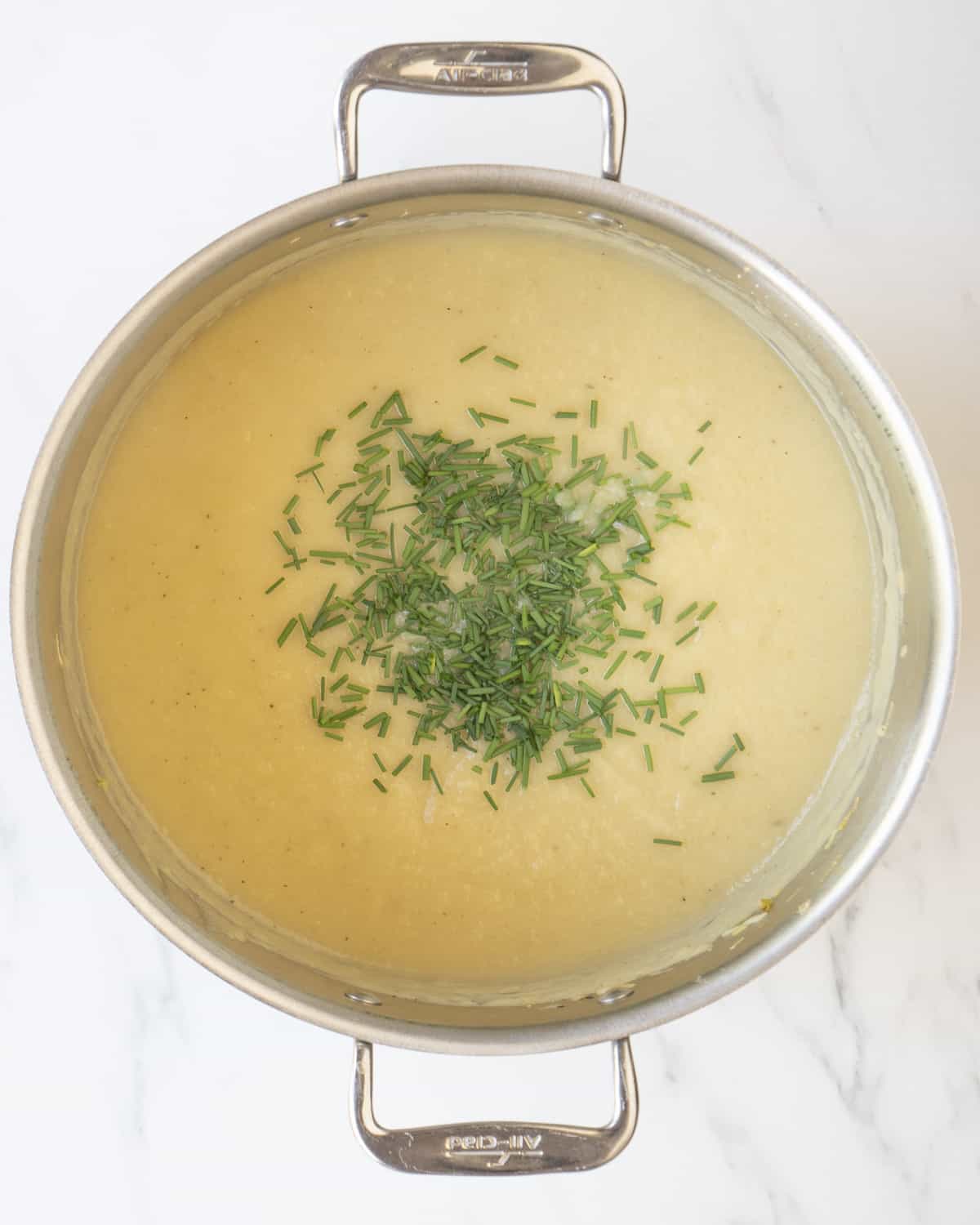 A dutch oven with blended potato leek soup topped with chopped chives on a white countertop. 