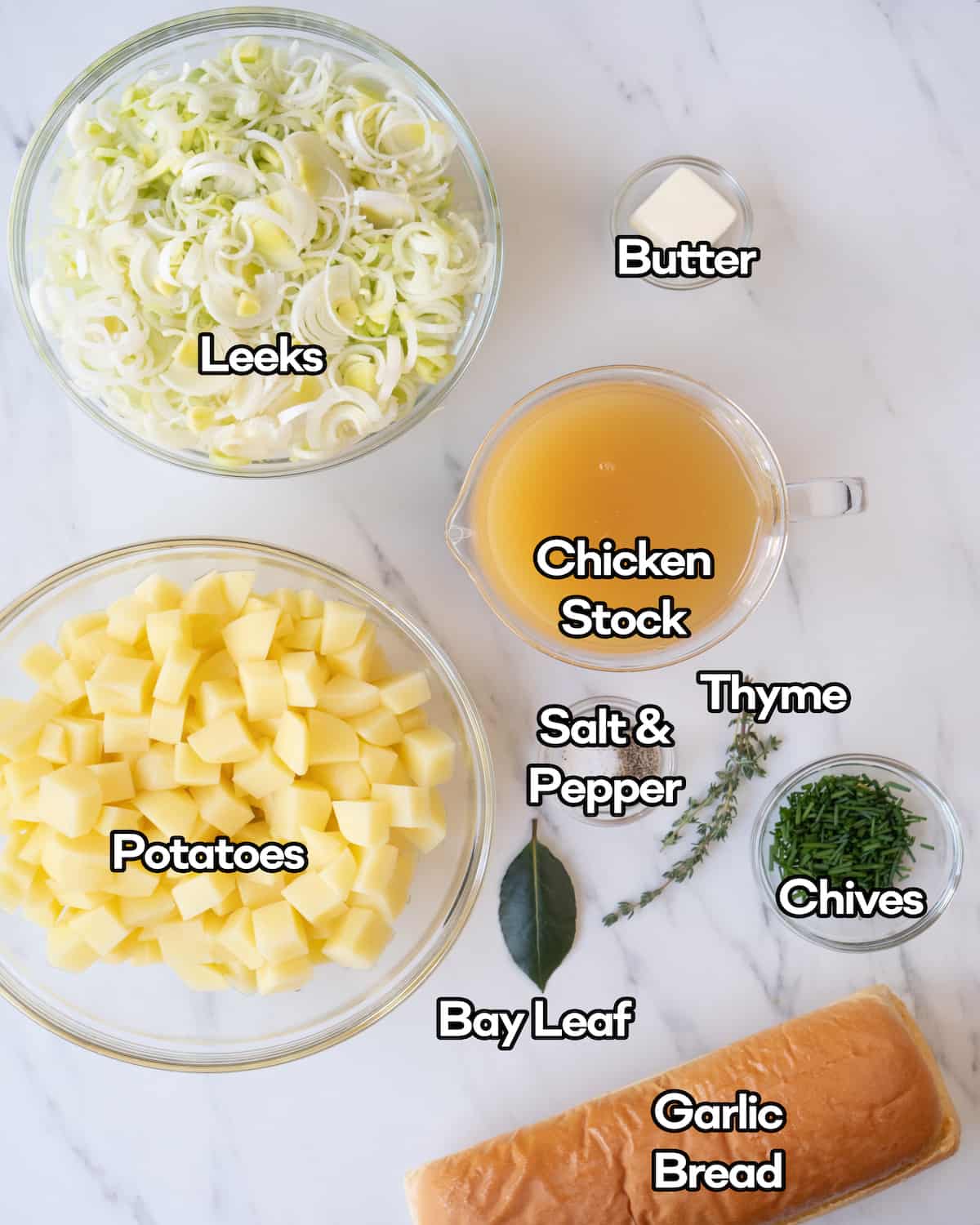 Ingredient shot of each individual ingredient in clear bowls including leeks, butter, potatoes, chicken stock, salt and pepper, chives, thyme, bay leaf, and garlic bread.