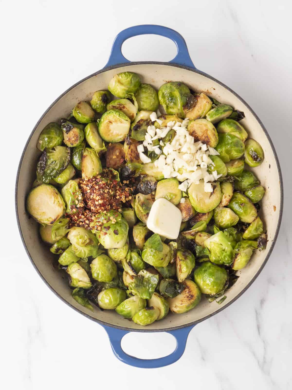 Large blue dutch oven with sautéed brussels sprouts topped with chopped garlic, a tablespoon of butter and red pepper flakes.