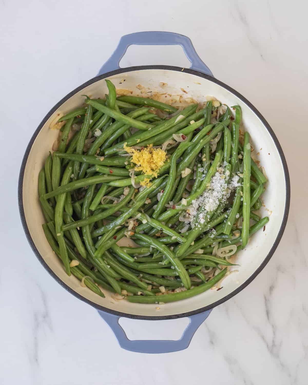 Shallot, garlic cloves, red pepper flakes, green beans, lemon zest, and flaky salt in a blue skillet on a white countertop.