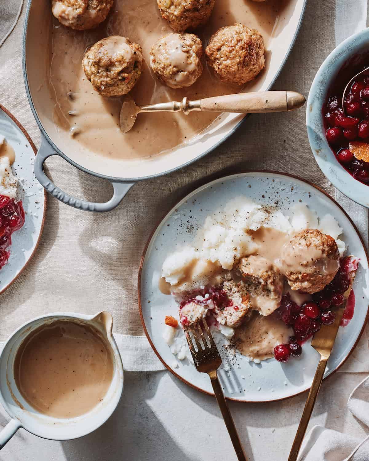 Half view of oval shallow dish with thanksgiving turkey meatballs in gravy with a big serving fork, placed on a table layered with a cloth, a bowl with cranberry sauce peeking from the side, a gravy boat with more gravy and a plate of mashed potato, meatballs and cranberry sauce.