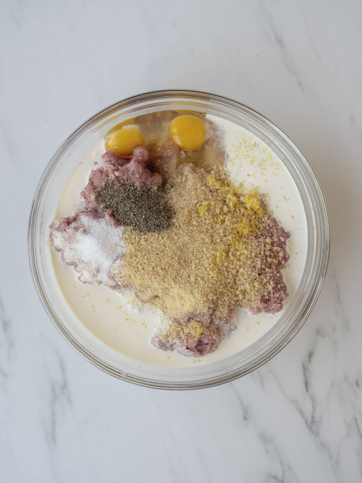 A large glass mixing bowl with ground turkey, ground pork, breadcrumbs, lemon zest, heavy cream, eggs, and salt and pepper.