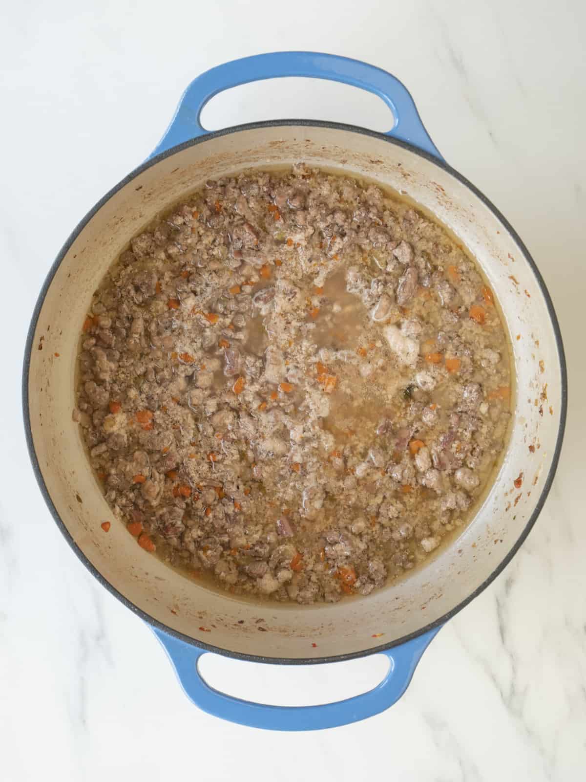 A large blue dutch oven with the cooked vegetables and meats cooking, and chicken stock added.