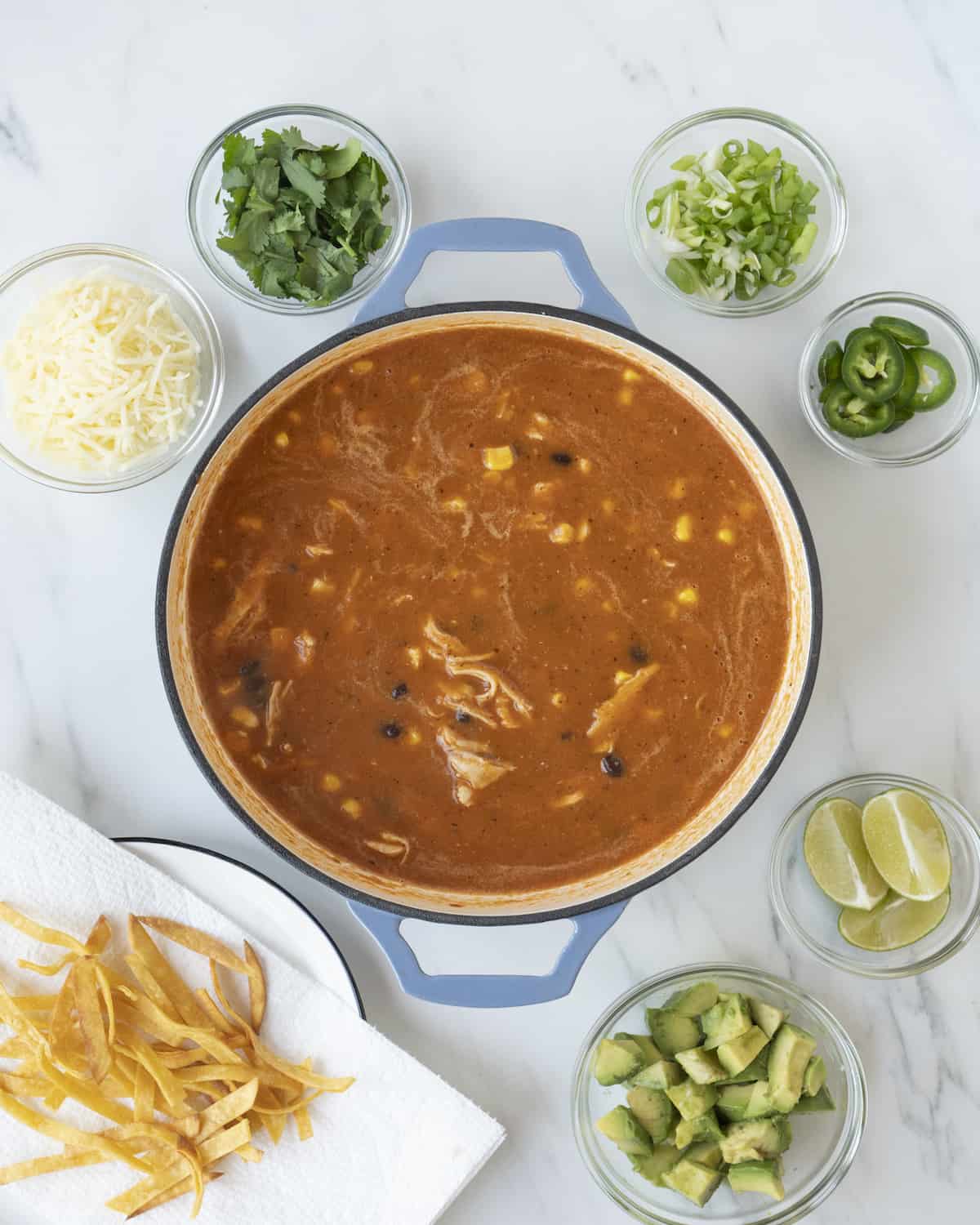 A blue saucepan with completed chicken tortilla soup recipe surrounded by clear bowls of ingredient toppings such as cheese and cilantro in top left corner, a plate of fried tortilla strips in lower left corner, clear bowls of green onion and jalapeños in top right corner, and clear bowls of 3 lime wedges and cubed avocado in lower right corner on a white countertop.