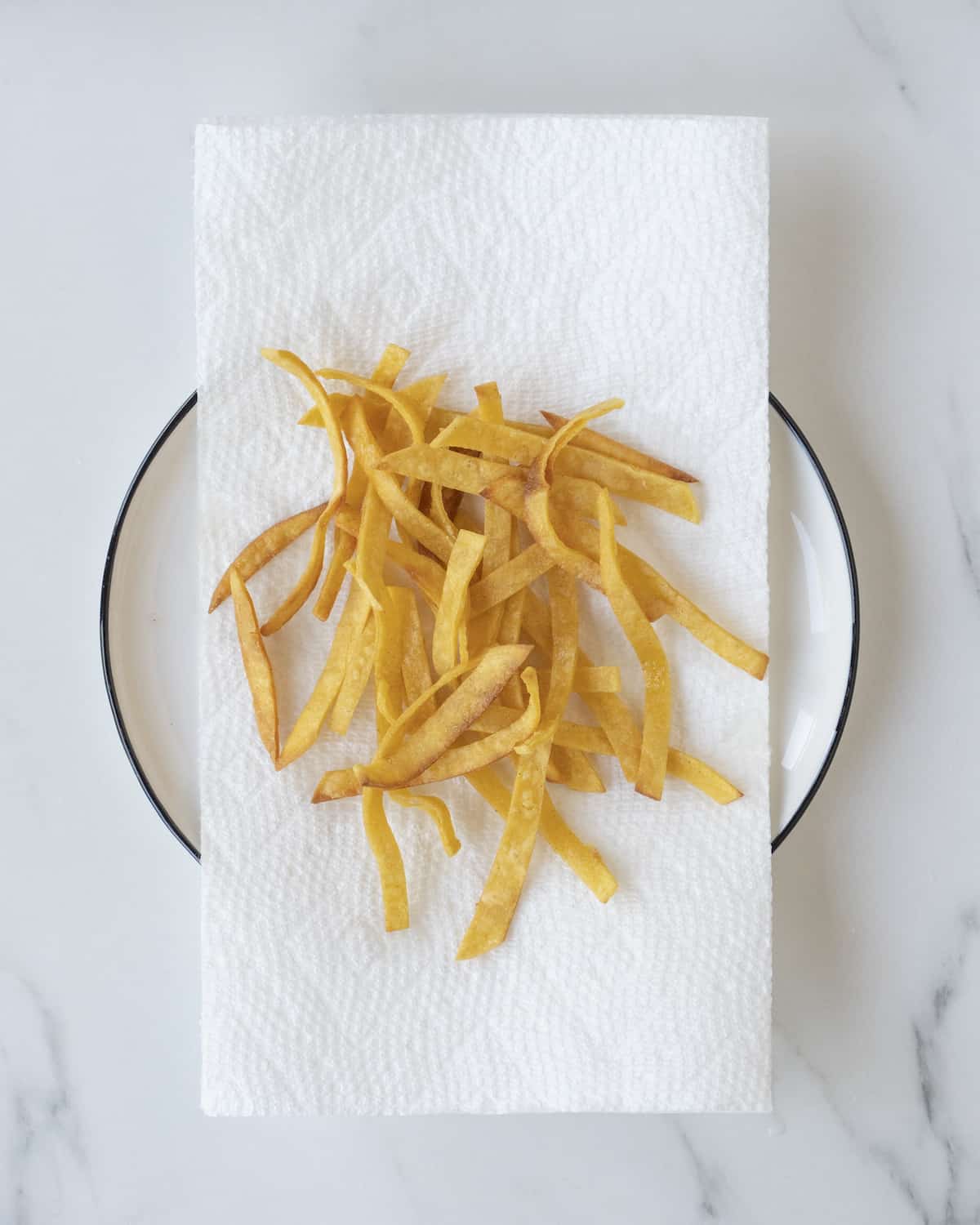 A ceramic plate with fried tortilla strips and a paper towel beneath them on a white countertop. 