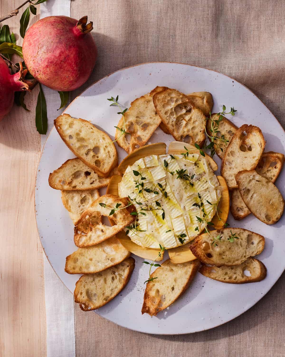 A white plate with Baked Camembert on the center, surrounded by toasted crostinis and topped with thyme, with pomegranates next to the plate.