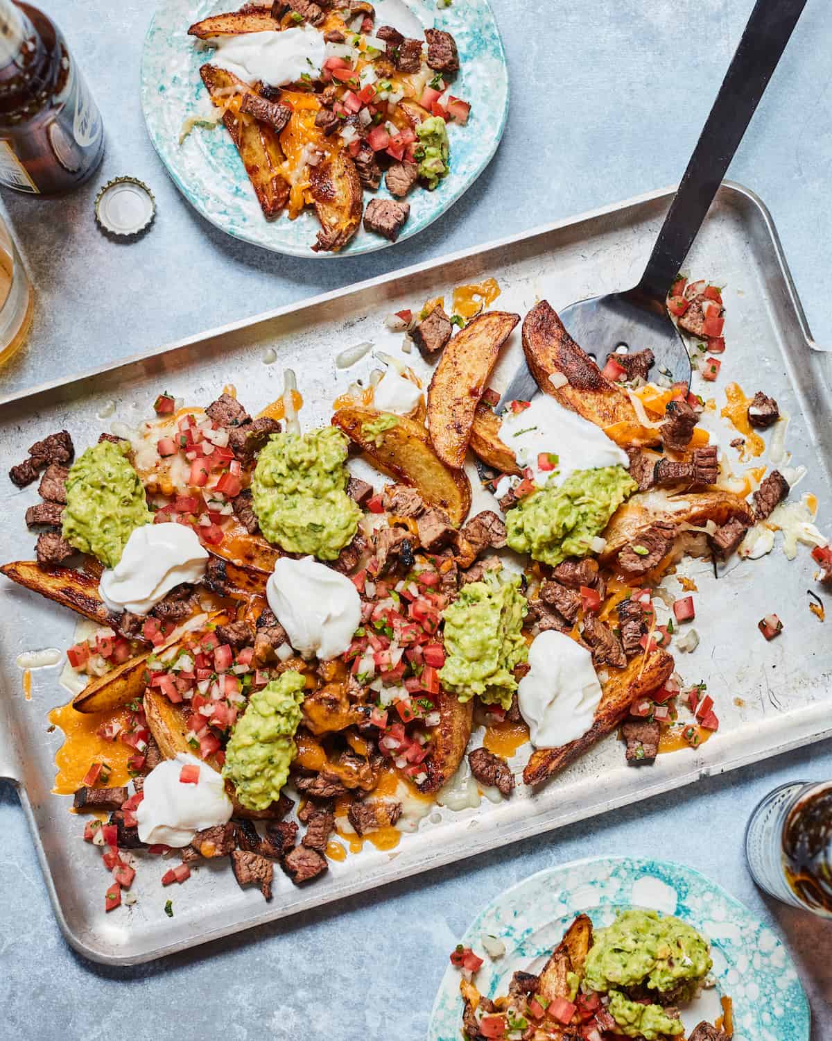 Sheet pan with carne asada nacho fries topped with sour cream and guacamole with a black metal spatula on a blue countertop with a blue ceramic plate of nacho fries in top left corner and bottom right corner as well as an open bottle of beer in the top left corner. 