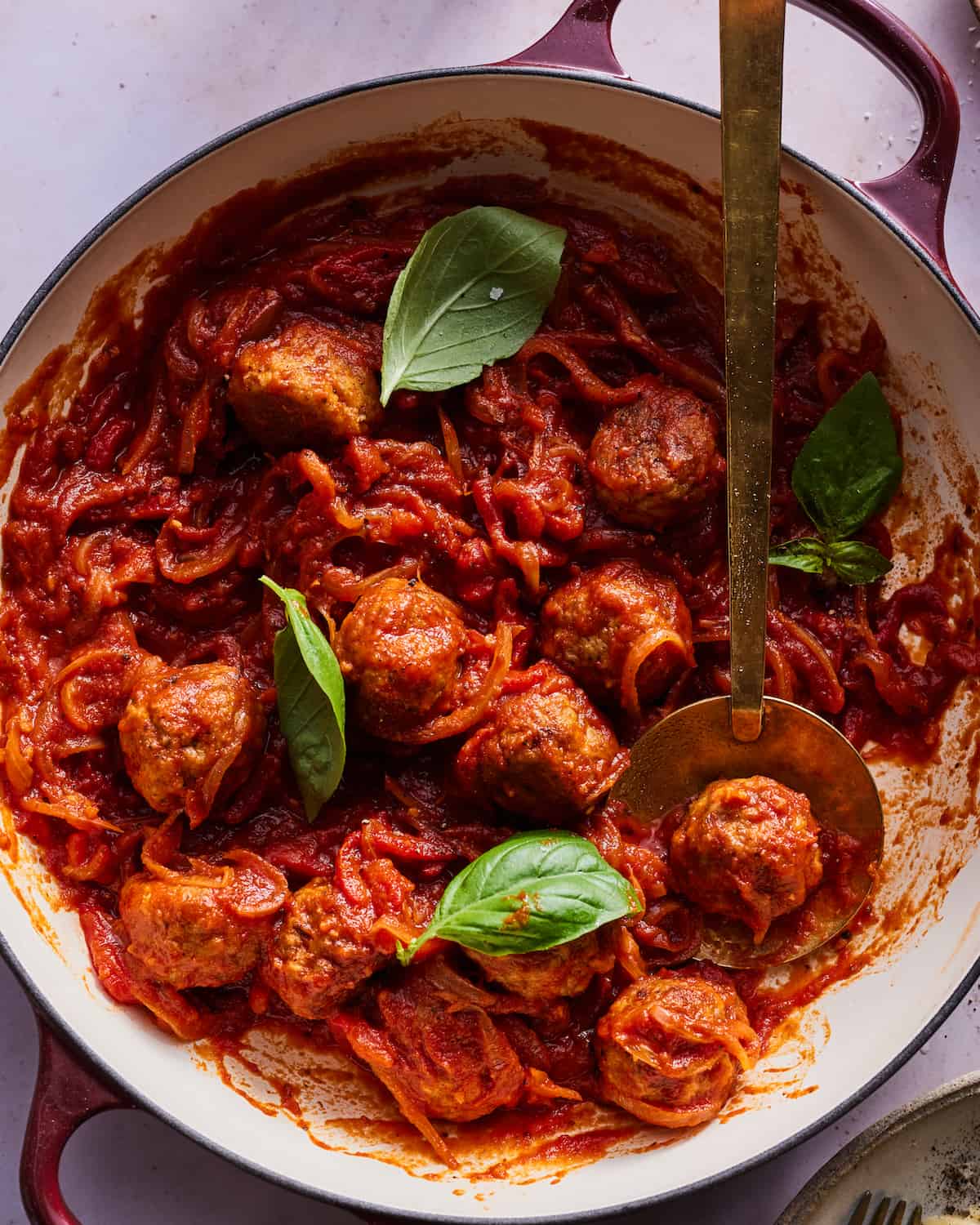 A braiser with chicken meatballs in red sauce with a garnish of basil on a purple table.
