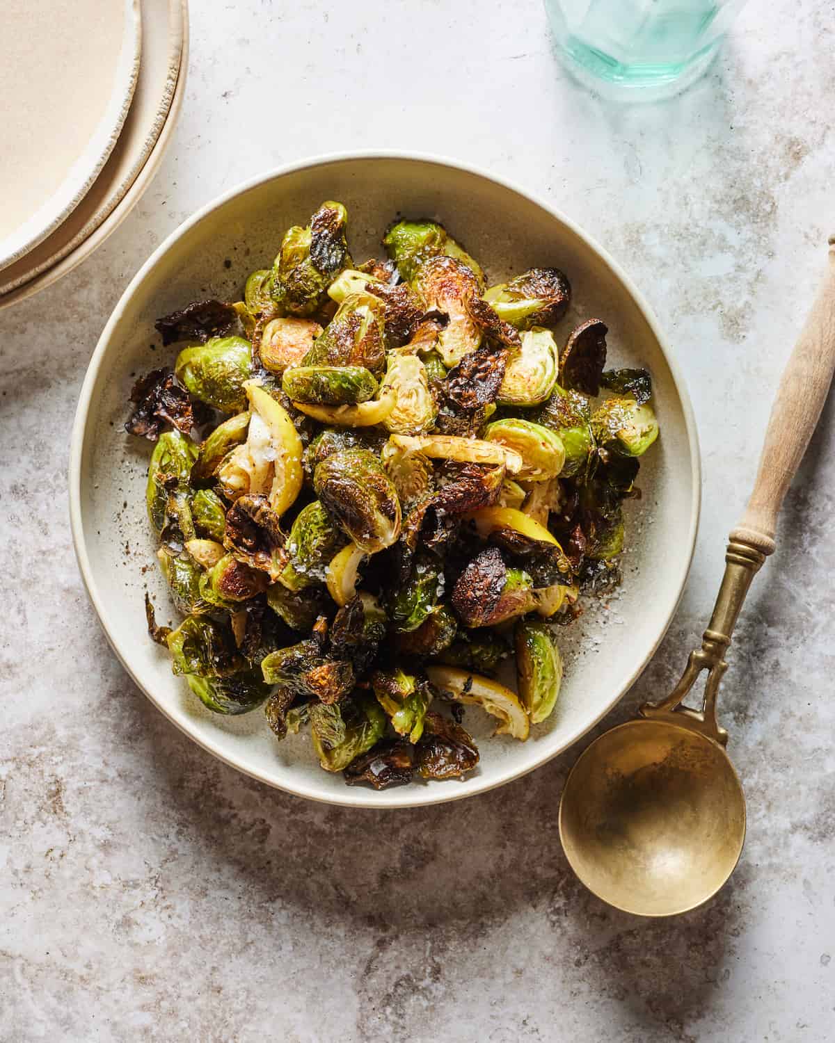A round flat dish with crispy roasted brussels sprouts, with a golden serving spoon on the side.