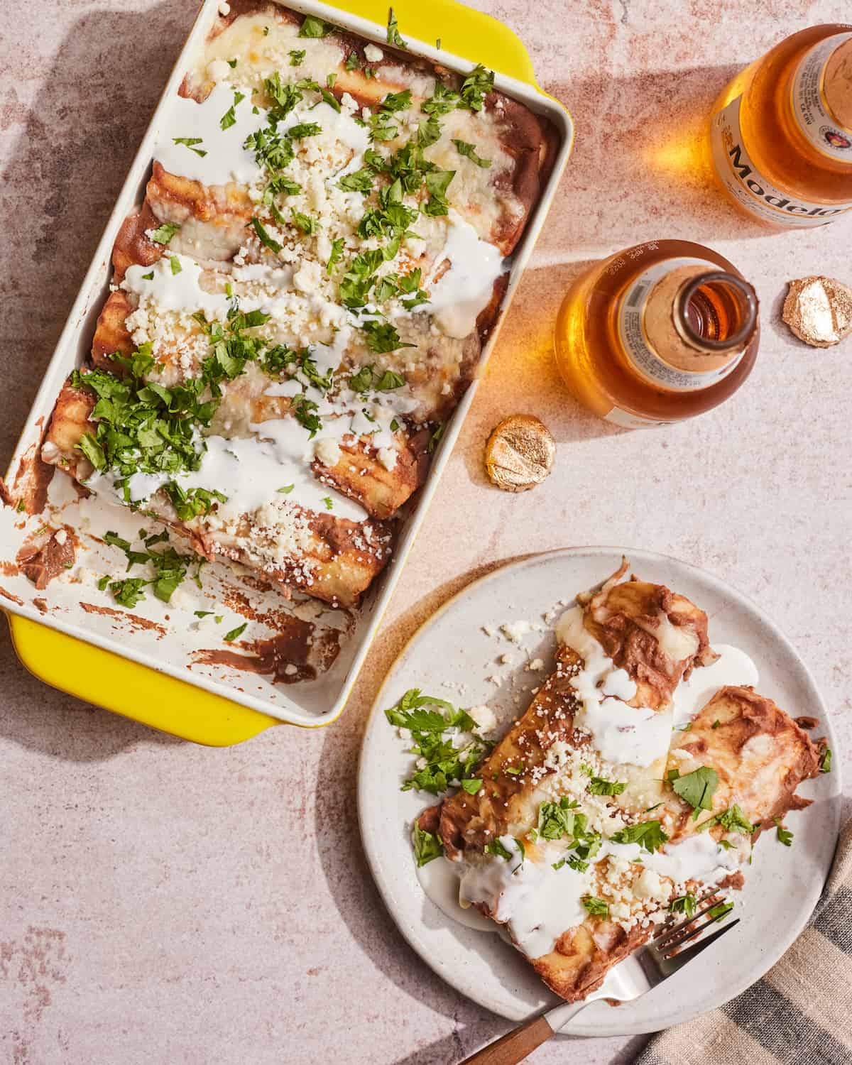A rectangular white baking dish with black bean puree enchiladas, also called Enfrijoladas, with a plate of the same dish on the side and two modelo beer bottles.